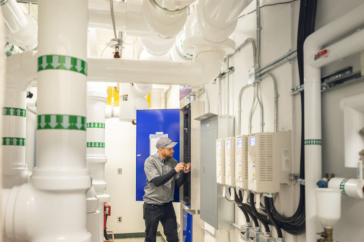 A Kimco technician in a grey Kimco cap and shirt adjusts wiring inside an open controls panel in a mechanical room, next to an electrical panel, a row of equipment drives and white insulated piping.