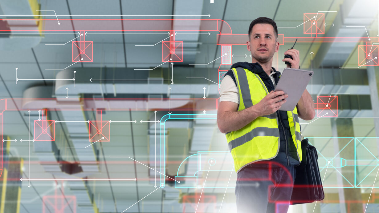 A field technician in a high-visibility vest holds a tablet and radio while reviewing building system diagrams overlaid on a commercial ceiling, representing real-time building automation work on site. 
