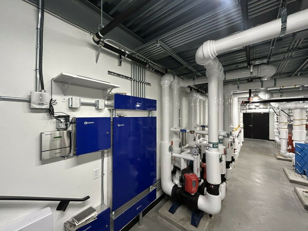 Mechanical room with two blue Kimco DDC control panels on the wall beside overhead piping and heating and chilled-water pump equipment.